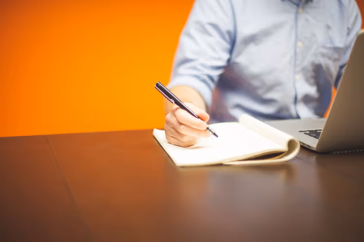 Person taking notes at a desk next to a laptop