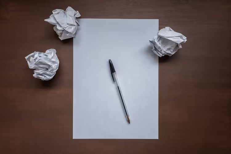 Leeres Blatt, Stift und zerknüllte Papierkugeln auf einem Schreibtisch als Symbol für Schreibblockade