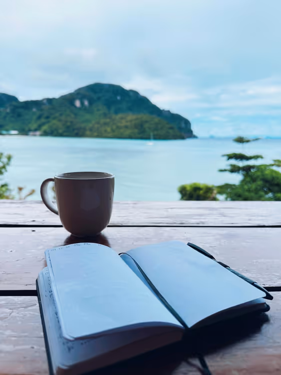 Journal ouvert et tasse de café sur une table en bord de mer, séance d'écriture matinale en voyage