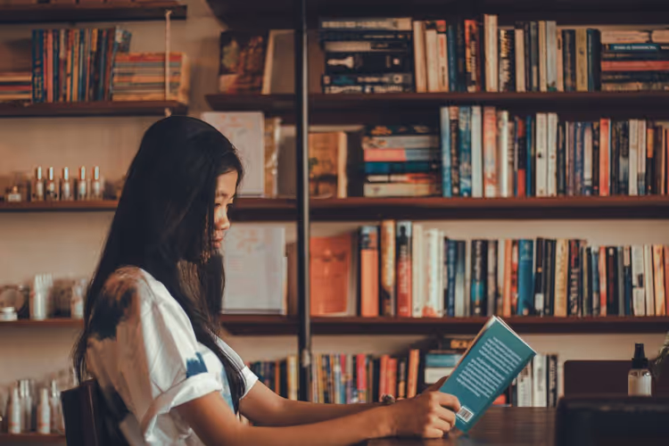 A girl reads a book in a library next to the shelves